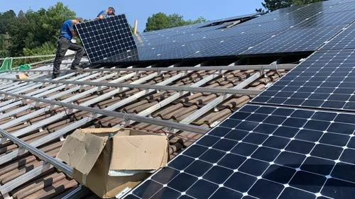 Zwei Monteure setzen ein Solarmodul auf die Aluminiumschienen eines Ziegeldachs, daneben liegen bereits fertig montierte Module